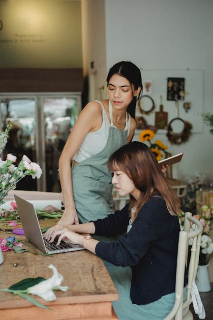 Two women in a flower shop, one standing and wearing a light blue apron and white tank top, observing a laptop screen, while the other seated woman, with shoulder-length brown hair and wearing a dark