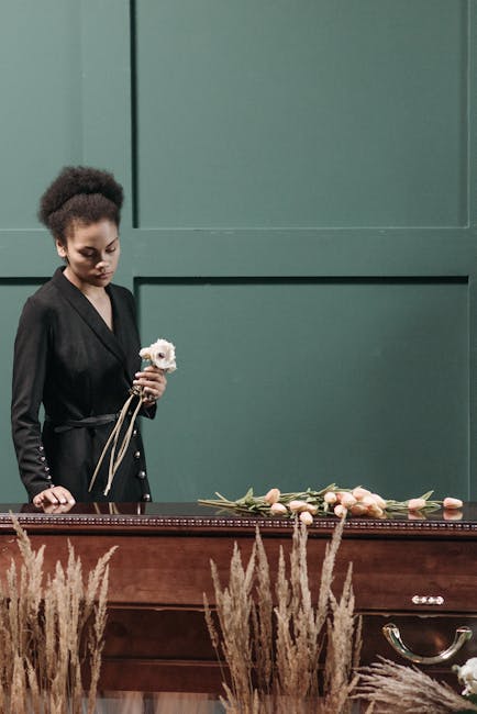 A young woman with curly dark hair, dressed in an elegant black outfit, stands at a wooden casket with a solemn expression. She holds a white flower, possibly a rose or lily, in her hand. In front of 