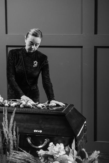 A woman arranging a floral display on top of a dark, polished coffin during a funeral service. She is dressed in a black, long-sleeved, textured outfit with a brooch on her chest, focused on carefully