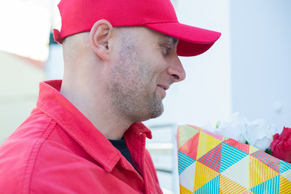 A man wearing a bright red baseball cap and matching red jacket is holding a colorful floral gift bag filled with white and red flowers. The man is facing to the right with a gentle smile, and the bac