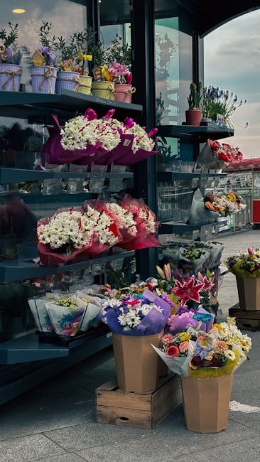 A display of flower arrangements and bouquets on black metal shelves inside a floral shop near a large window with a view of the city and Euston Station. The arrangements include white, pink, purple,
